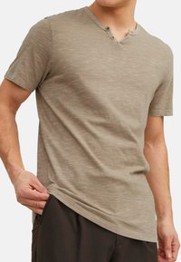 Beige kortærmet t-shirt med en tekstureret overflade, der har en V-halsudskæring og to knapper ved kraven, kombineret med mørke shorts.