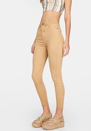 Beige hoge taille skinny jeans met een soepele textuur, voorzien van een sluiting met een knoop aan de voorkant en subtiele stikdetails. Gecombineerd met tan wedge sandalen.