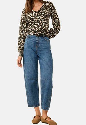 Vrouw die een lange mouwen top in luipaardprint draagt, met een high-waisted blauwe spijkerbroek en een zwarte riem, en bruine platte schoenen, staand met één hand op haar heup.