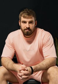 Haut rose clair à manches courtes avec un col rond et un logo brodé. Le tissu semble doux ; le modèle est assis, les bras croisés et porte un bracelet noir.