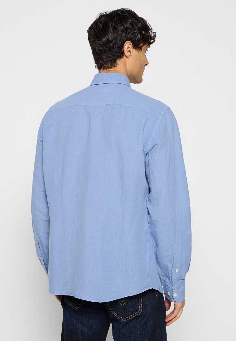 Camisa de manga larga azul claro hecha de algodón, con cuello abotonado, aberturas laterales y dobladillo recto. La vista trasera muestra costuras mínimas.