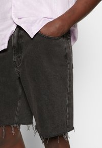 Shorts en denim noir avec des ourlets effilochés, design à cinq poches et coutures contrastantes. Portés avec une chemise à motifs lavande clair.