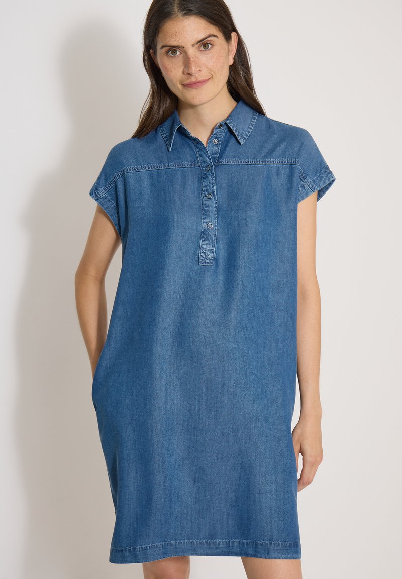 Femme aux cheveux bruns portant une robe en jean bleue longueur genoux avec des manches courtes et un col boutonné, debout devant un fond uni.