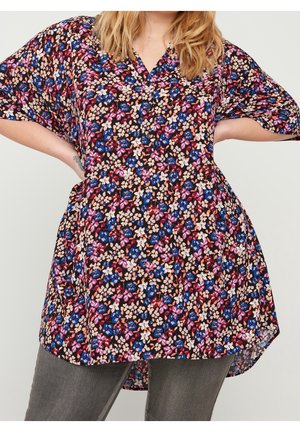 Femme portant une tunique fleurie ample avec des fleurs roses, bleues et beiges sur un pantalon gris, mains sur les hanches, cheveux blonds et ondulés.