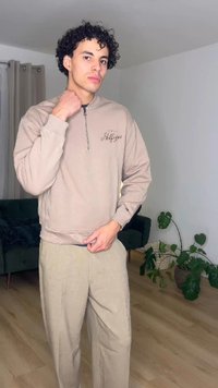 Νεαρός άνδρας που ρυθμίζει το γιακά ενός μπεζ φούτερ με φερμουάρ, στεκόμενος μέσα σε σπίτι κοντά σε ένα πράσινο καναπέ και φυλλώδη φυτά σε γλάστρες πάνω σε ξύλινο δάπεδο.