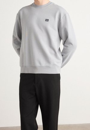 Mann trägt ein hellgraues Sweatshirt mit langen Ärmeln und kleinem schwarzem Logo auf der Brust sowie schwarze Hose, steht mit den Händen hinter dem Rücken.