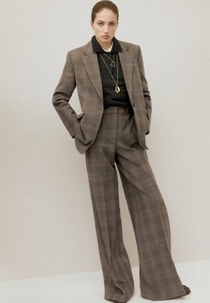 Traje de cuadros marrón con pantalones de pierna ancha. La chaqueta tiene un ajuste relajado con un sutil corte, usada sobre un suéter negro y acentos de collar dorado.