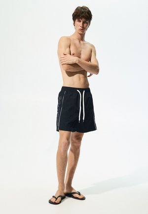 Schwarze Badehosen mit weißem Kordelzug und seitlichem grafischen Design. Model trägt schwarze Flip-Flops und steht mit verschränkten Armen.