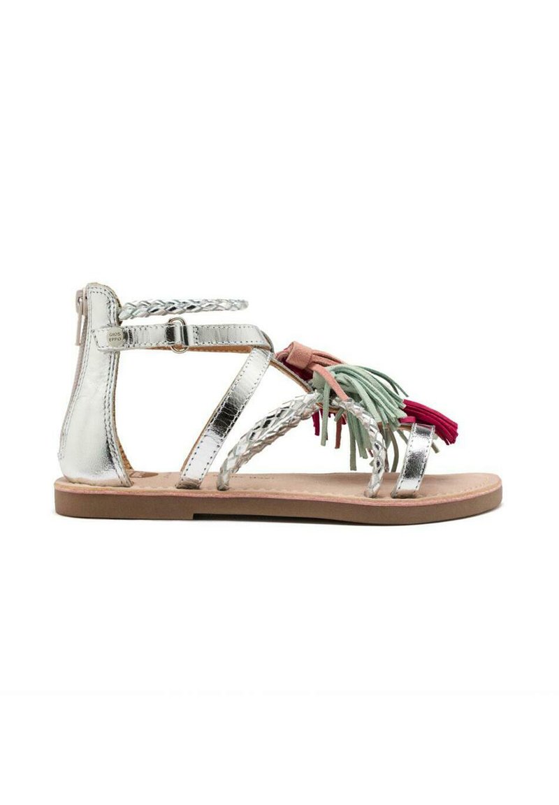 Sandalias metálicas plateadas con correas trenzadas y adornos de borlas coloridas en verde y rojo. Suela plana, diseño abierto y correa de tobillo ajustable.