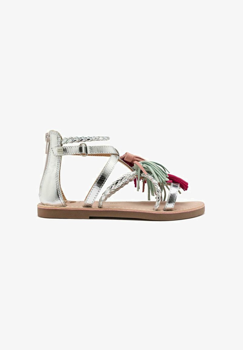 Sandalias metálicas plateadas con correas trenzadas y adornos de borlas coloridas en verde y rojo. Suela plana, diseño abierto y correa de tobillo ajustable.
