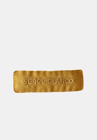 Étiquette en tissu dor é avec du texte en relief indiquant "SERGE BLANCO." Forme rectangulaire, texture lisse et bords renforcés.
