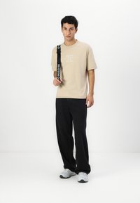 Beige bomull t-skjorte med trykt logo, kombinert med svart løstsittende bukser og grå joggesko, stående mot en ensfarget bakgrunn.