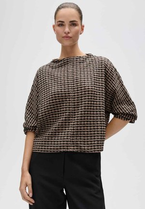 Femme aux cheveux foncés attachés en arrière, portant un haut ample à motifs avec manches retroussées et pantalon noir, debout devant un fond uni.