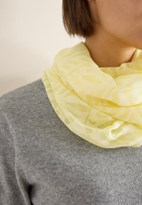 Écharpe jaune clair en tissu transparent et texturé présentant un motif abstrait subtil, drapée autour du cou sur un pull gris.