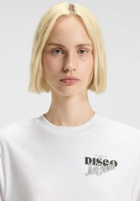 Valkoinen puuvillateeppari, jossa on pyöreä kaula-aukko ja musta graafinen printti, jossa lukee "DISCO Melodrama" vasemmassa rinnassa.