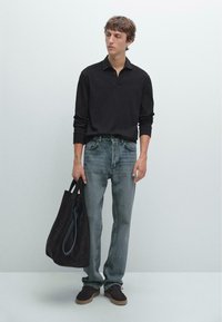 Schwarzes Langarm-Polo, ausgebleichte blaue Jeans und braune Wildledertasche; das Modell trägt dunkle Schuhe; neutraler Hintergrund.