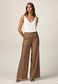 Bluză albă fără mâneci cu decolteu în V, asortată cu pantaloni maro, cu talie înaltă și pantaloni larghi, având un model texturat și buzunare frontale. Sandale negre.