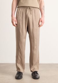 Pantalons beiges à taille élastique avec cordon de serrage, présentant une coupe droite et des coutures minimalistes, associés à des chaussures noires sur un fond neutre.