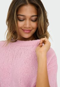 Lichtroze gebreide trui met een losse pasvorm, geribbelde halslijn en een gestructureerd patroon. Model houdt de halslijn vast met een ontspannen uitstraling.