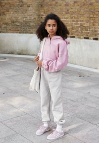 Pink hooded sweatshirt with elastic cuffs, gray tapered sweatpants, and pink sneakers. Carrying a light beige tote bag. Textured background.