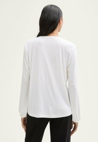 Weißes Langarmoberteil mit Rundhalsausschnitt und gerafften Rippärmeln, aus einem weichen, leichten Stoff, kombiniert mit schwarzen Hosen.