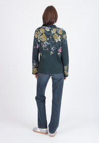 Maglione verde scuro con motivo floreale giallo, rosa e bianco. Indossato con jeans blu e scarpe chiare. Vista da dietro, sfondo semplice.
