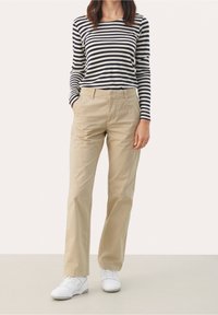 Žena v béžových rovně střižených kalhotách, černobílém pruhovaném tričku s dlouhým rukávem, s rukama v kapsách a bílými teniskami.