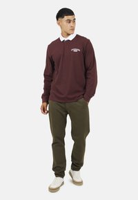 Polo a maniche lunghe color bordeaux con colletto bianco, logo sul petto, abbinato a pantaloni verde oliva e scarpe da ginnastica bianche. Design semplice, vestibilità casual.