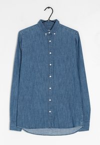 Camisa de mezclilla en azul medio, de mangas largas, con cuello botón, que presenta una tapeta delantera con botones blancos y un sutil logotipo en el pecho.