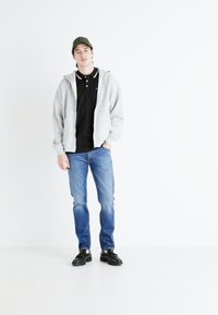 Gray zip-up hoodie over a black polo shirt, blue denim jeans, and black sneakers. Accessories include a green cap. Plain white background.