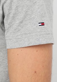 T-shirt en coton gris à manches courtes, avec un petit logo de drapeau brodé en rouge, blanc et bleu sur la manche.