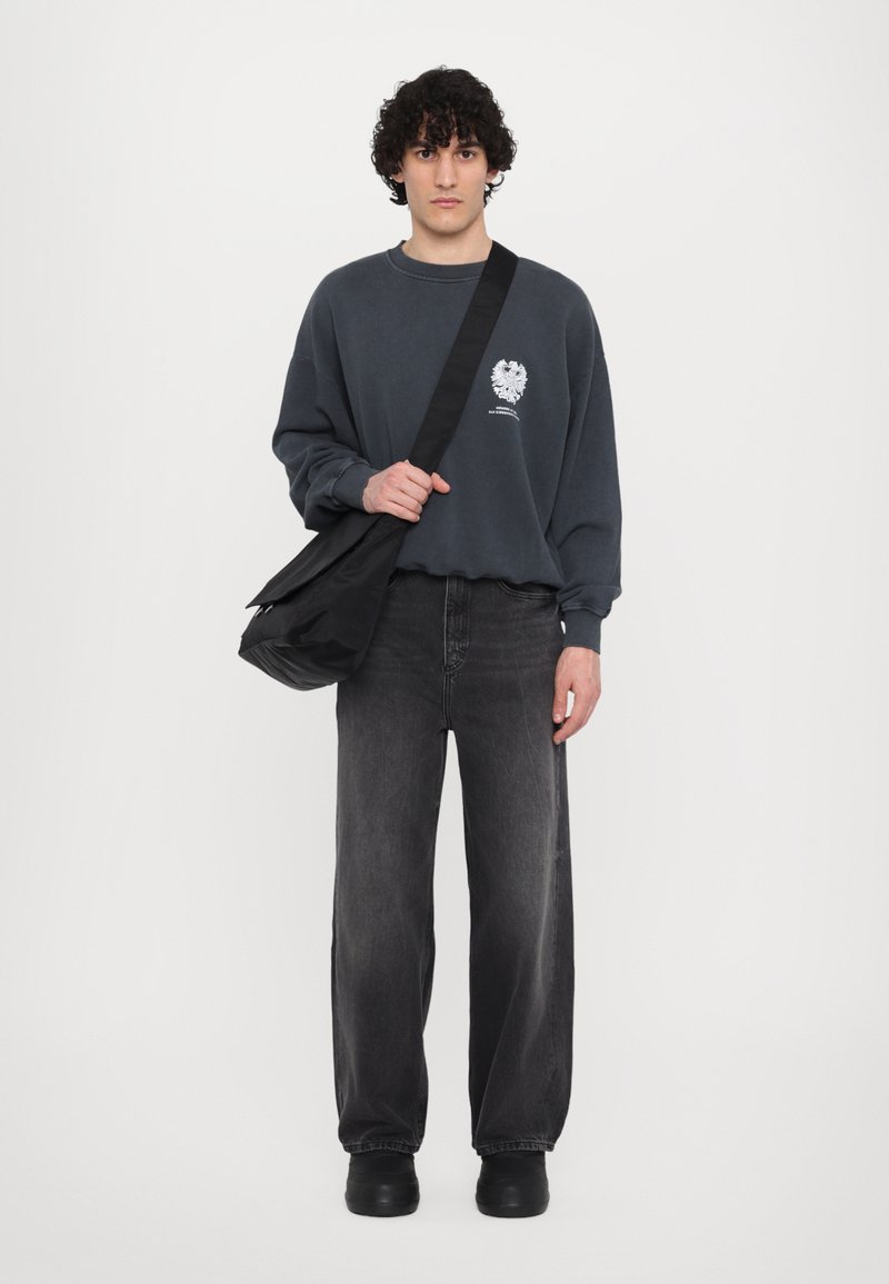 Sweatshirt gris avec logo, jean noir à jambes larges et sac à main noir. Le mannequin porte des chaussures à plateforme noires. Design simple et décontracté.