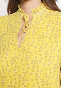 Blusa amarilla con estampado floral, con un cuello con volantes y un cierre con lazo en el escote, hecha de un tejido ligero.
