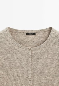 Cardigan beige en maille côtelée avec col rond et six petits boutons ronds au centre devant.