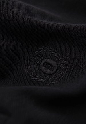 Gros plan sur un tissu noir texturé présentant un logo brodé circulaire avec une couronne de laurier et une initiale au centre, suggérant une marque.