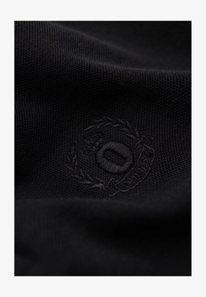 Nahaufnahme von strukturiertem schwarzem Stoff mit einem runden gestickten Logo, das einen Lorbeerkranz und ein Initial in der Mitte zeigt und auf eine Marke hindeutet.