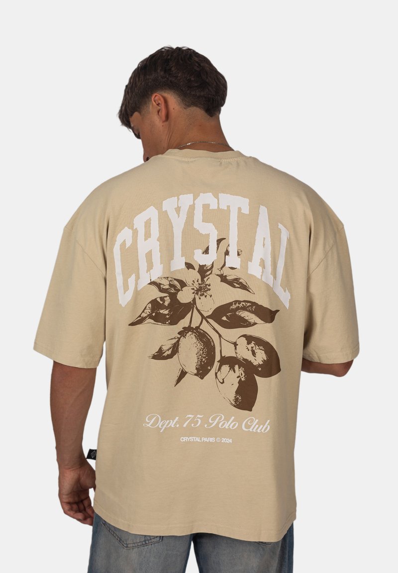 Béžové oversized tričko se velkým bílým textem "CRYSTAL" a hnědým květinovým designem s citrony na zádech. Krátké rukávy, volný střih.
