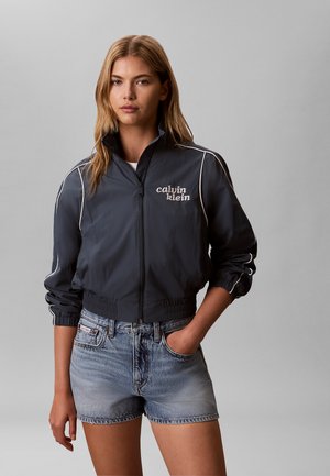 Kobieta ubrana w ciemną kurtkę na zamek z białymi lamówkami i logo Calvin Klein, w połączeniu z jasnymi szortami dżinsowymi, stojąca na tle jednolitego szarego tła.