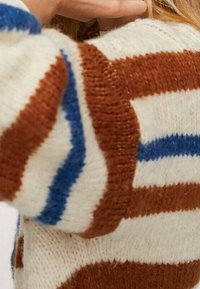 Sweter dziergany z szerokimi poziomymi paskami w kolorach brązowym, niebieskim i kremowym na osobie z długimi blond włosami.