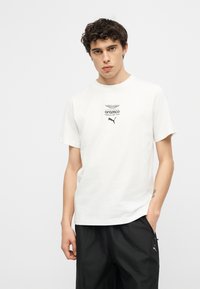 Νεαρός άνδρας με σγουρά μαλλιά, φορώντας λευκό t-shirt της ομάδας Aramco Formula One της Puma και μαύρο π αντελόνι Puma, στέκεται μπροστά σε απλό φόντο.