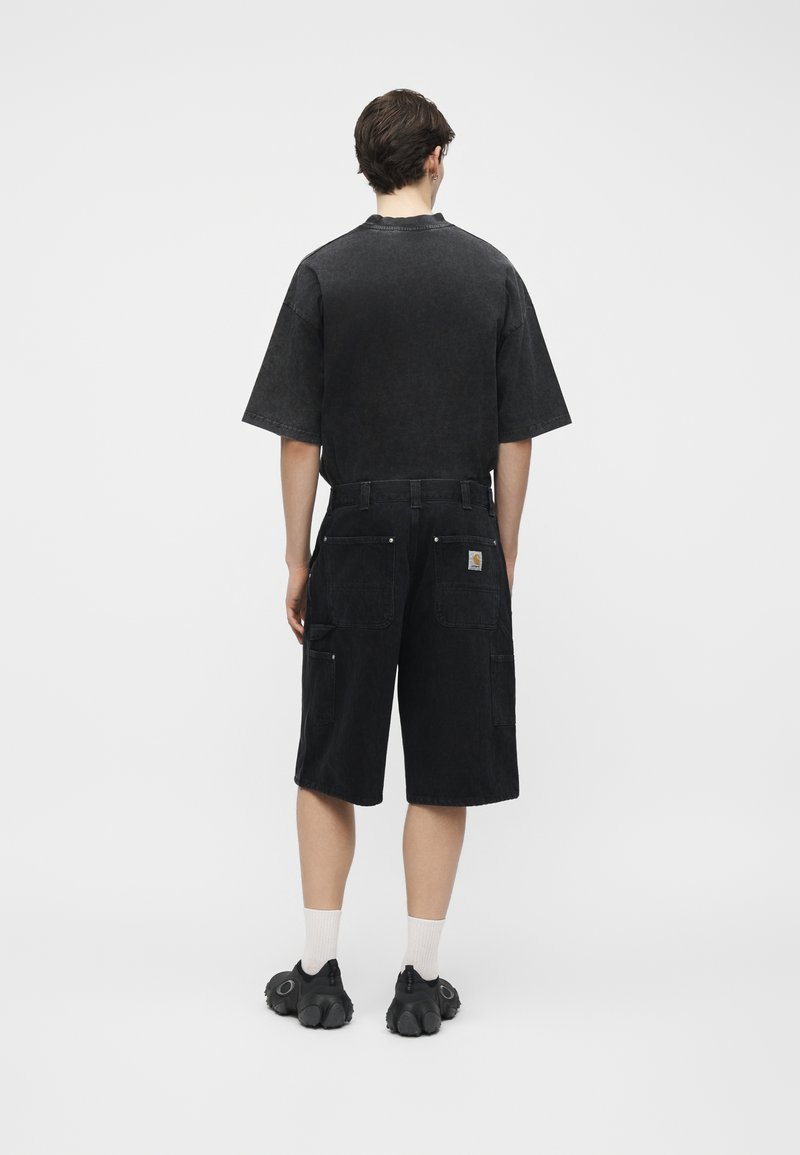 Mann, der von hinten zu sehen ist, trägt ein schwarzes übergroßes T-Shirt, schwarze knielange Shorts mit Taschen, weiße Socken und schwarze Schuhe vor einem einfarbigen Hintergrund.