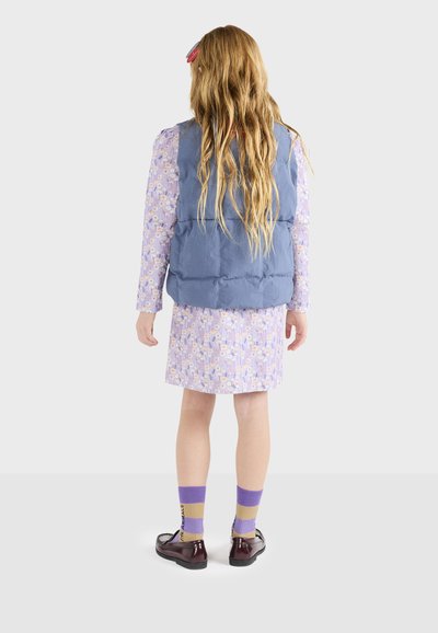 Blå puffy vest over en mønstret lilla kjole, parret med lilla og gyldne stribede sokker og mørkebrune sko. Langt bølget hår synligt.
