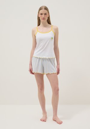 Jeune femme debout pieds nus portant un caraco blanc avec des bordures jaunes et un short rayé bleu et blanc avec des bords jaunes.