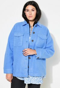 Chaqueta de forro polar azul con dos bolsillos frontales, cierre de botones y un corte relajado. Llevada sobre una camisa de rayas azul y blanca. Pantalones negros mostrados.
