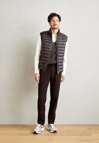 Zwarte puffer vest met horizontale stiksels, gedragen over een grijze v-hals trui, zwarte broek en grijs-witte sneakers op een houten vloer.