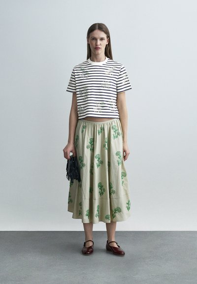 Γυναίκα στέκεται φορώντας ένα λευκό και ναυτικό ριγωτό κοντό πουκάμισο με λουλουδάτα σχέδια, μια μπεζ midi φούστα με πράσινα λουλουδάτα μοτίβα, κρατώντας μια μαύρη τσάντα.