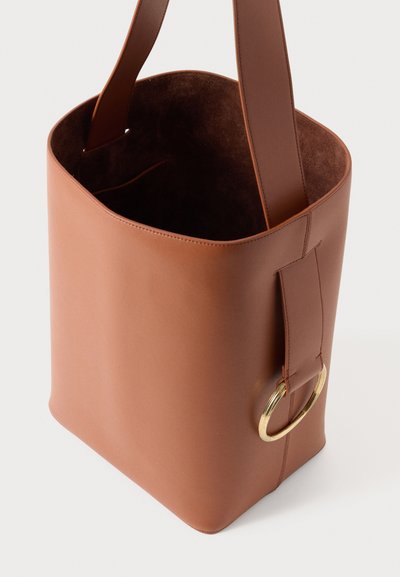 Hnedá kožená taška typu tote s hladkým povrchom, vybavená širokým plochým remienkom a zlatým kovovým krúžkom ako ozdobou. Vnútro má semišovú textúru.