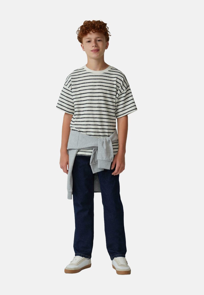 Chemise à rayures à manches courtes en crème et gris, associée à un jean bleu foncé. Pull gris noué à la taille et baskets blanches avec semelles brunes.