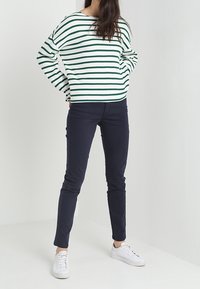 Nainen seisoo kädet taskuissa, päällään valkoiselle ja vihreälle raidallinen neule, tummansiniset housut ja valkoiset lenkkarit tasaisella taustalla.