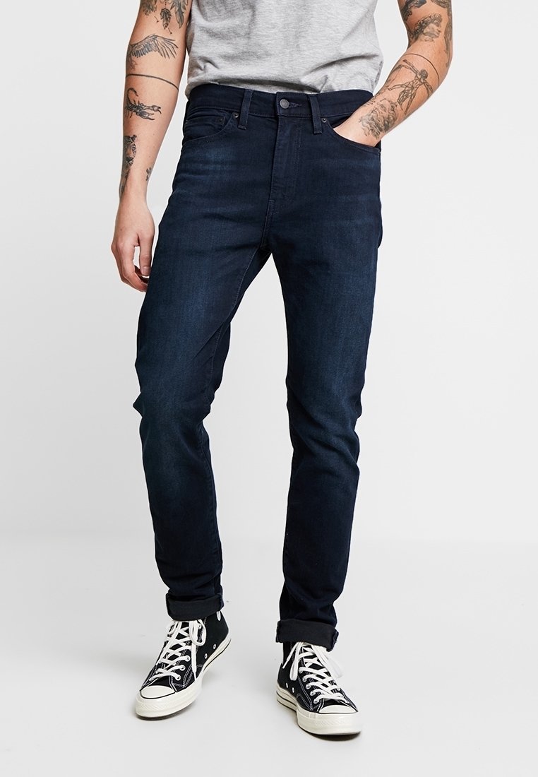Jean bleu foncé en denim, design slim-fit, comportant cinq poches et des coutures discrètes. Portés avec des baskets noires et blanches.